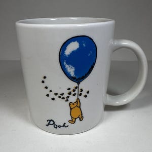 Pode incluir: Caneca de cerâmica branca com um balão azul, Winnie the Pooh segurando-se nele, e o texto "Pooh" na parte inferior.