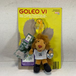 Könnte beinhalten: Ein Goleo VI Löwen-Maskottchen der FIFA Fussball-Weltmeisterschaft 2006 in Deutschland, befestigt an einer gelben Karte. Der Löwe trägt ein weißes Trikot mit der Nummer 06 und schwarze Schuhe. Die Karte hat Text auf Deutsch und Englisch.