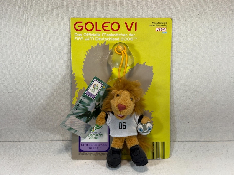May include: A plush Goleo VI lion mascot from the 2006 FIFA World Cup Germany, attached to a yellow backing card. The lion wears a white shirt with the number 06 and black shoes. The card has text in German and English.