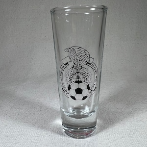 Puede incluir: Un vaso de chupito de vidrio transparente con un logotipo en blanco y negro de la Federación Mexicana de Fútbol. El logotipo presenta un águila con un balón de fútbol en sus garras.