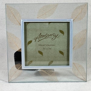 Könnte beinhalten: Ein silberner Bilderrahmen mit einer Glasfront und einem dekorativen Design aus gepressten Blättern. Der Rahmen ist 8,9 cm x 8,9 cm groß und trägt den Text "Accessorize" und "Marcel Schurman" auf der Vorderseite.