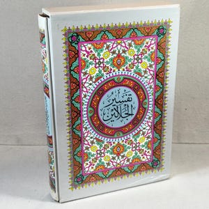 Op de afbeelding: Een hardcover boek met een witte kaft met sierlijke, kleurrijke bloemen- en geometrische ontwerpen. De kaft bevat een centraal medaillon met Arabisch schrift. De rug van het boek is ook zichtbaar, met tekst en ontwerp.