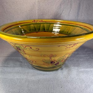 May include: A wide, shallow ceramic bowl featuring a warm yellow base color with hand-painted green and dark red vine and grape motifs. The interior rim is accented with a deep green band, while the exterior displays swirling decorative lines.