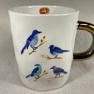May include: White ceramic mug with a gold handle, featuring hand-painted blue birds perched on branches. The mug has a slightly textured surface and a small, round, brown sticker with the number 4.