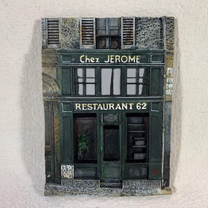 May include: A green building with a sign that reads "Chez Jerome Restaurant 62". The building has a stone facade and a wooden door. There are windows on the second floor and a sign that says "Ete Off Gaz" on the right side of the building.