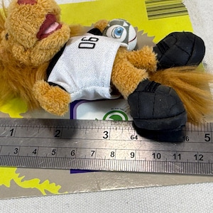 May include: A plush lion toy wearing a white jersey with the number 06, black shorts, and black shoes. The lion is holding a small soccer ball. The toy is approximately 4 inches long.
