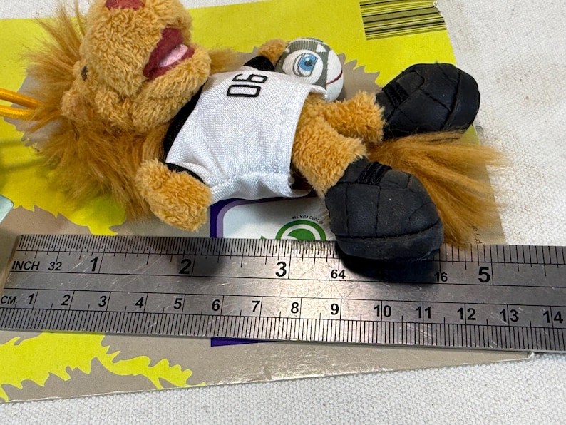 May include: A plush lion toy wearing a white jersey with the number 06, black shorts, and black shoes. The lion is holding a small soccer ball. The toy is approximately 4 inches long.