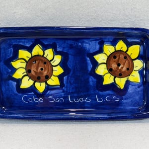 Puede incluir: Una bandeja rectangular de cerámica azul con dos girasoles amarillos pintados en ella. La bandeja tiene las palabras "Cabo San Lucas b.c.s." escritas en blanco en la parte inferior.