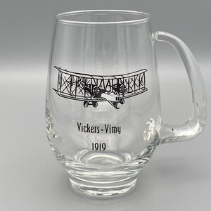 Könnte beinhalten: Klarer Bierkrug aus Glas mit gebogenem Griff. Der Krug zeigt eine schwarze Illustration eines Doppeldeckers und den Text "Vickers-Vimy 1919". Der Krug hat einen abgerundeten Boden und eine breite Öffnung.