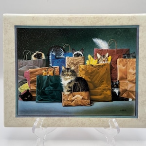May include: A rectangular ceramic tile featuring a detailed illustration of multiple cats peeking out from various colored paper shopping bags, including brown, blue, and green, set against a dark background with a light-colored border.