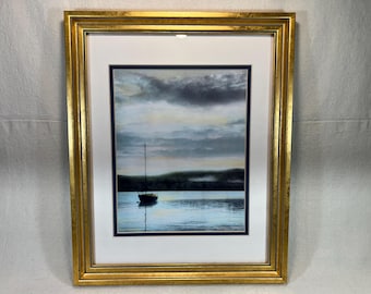 May include: A framed watercolor painting of a sailboat on a calm body of water. The sky is a mix of light blue and gray with fluffy clouds. The water is a light blue and reflects the sky.