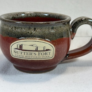 Puede incluir: Una taza de cerámica con forma redondeada y un asa. La taza presenta una combinación de colores rojo oscuro, marrón y verde. La parte delantera de la taza muestra una pegatina ovalada blanca con el texto "Sutter's Fort State Historic Park".