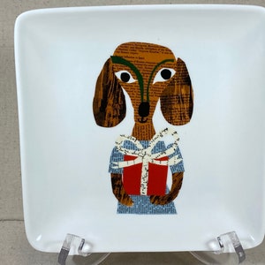 May include: A white ceramic square plate with a brown and white dachshund holding a red gift box with a white bow. The dachshund is made of paper collage.