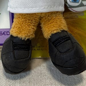 May include: Close-up of a plush toy's feet wearing black soccer cleats. The toy has tan fur legs and a white shirt. The image includes text that says "World Cup Germany 06" and "Licensed Product".
