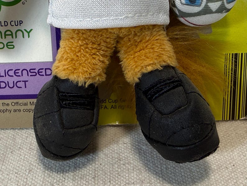 May include: Close-up of a plush toy's feet wearing black soccer cleats. The toy has tan fur legs and a white shirt. The image includes text that says "World Cup Germany 06" and "Licensed Product".