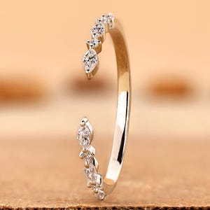 Pode incluir: Um anel aberto de ouro com uma série de pedras transparentes de corte marquise e redondo engastadas nas duas extremidades da banda. A banda de metal polido curva-se para criar uma abertura, com as pedras dispostas num padrão simétrico.