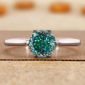 Op de afbeelding: Een zilveren ring met een grote, ronde, teal-kleurige edelsteen. De edelsteen heeft een facetachtig uiterlijk en de ringband is dun en gepolijst. De ring is geplaatst tegen een neutrale achtergrond.