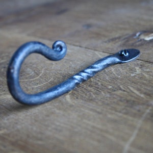Op de afbeelding: Een gedraaide metalen haak met een decoratieve krul aan de bovenkant. De haak is gemaakt van donkergrijs metaal en heeft een plat, afgerond uiteinde.