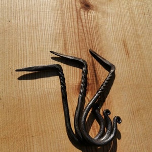May include: Three black metal hooks with a twisted design. The hooks are arranged in a stack and are on a light brown wooden surface.