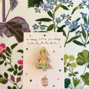 Könnte beinhalten: Ein hölzerner Weihnachtsbaum-Ansteck-Pin mit einem lächelnden Gesicht und bunten Punktdekorationen. Der Pin befindet sich auf einer weißen Karte mit dem Text "a happy little pin badge". Der Hintergrund ist ein Blumenmuster.