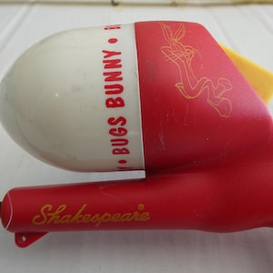 May include: A vintage red and white Bugs Bunny fishing rod reel with a yellow accent. The reel features the text "Bugs Bunny" and "Shakespeare" in gold lettering, along with a Bugs Bunny character outline.