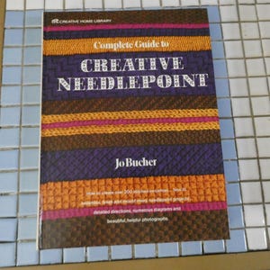May include: A book titled "Complete Guide to Creative Needlepoint" by Jo Bucher. The cover features a colorful, textured design with horizontal bands of orange, purple, brown, and pink. The book is part of the Creative Home Library series.
