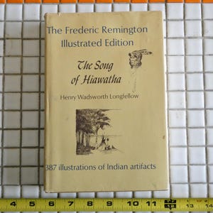 Könnte beinhalten: Ein Vintage-Buch mit dem Titel "The Song of Hiawatha" von Henry Wadsworth Longfellow, illustriert von Frederic Remington. Das Cover ist hellbraun mit schwarzem Text und Illustrationen. Das Buch enthält 387 Illustrationen indianischer Artefakte.