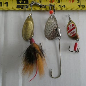 May include: Three fishing lures are shown against a yellow measuring tape. The lures include a gold-coloured spoon with a red and black feather tail, a silver hammered-metal spinner, and a red and white lure with a treble hook.