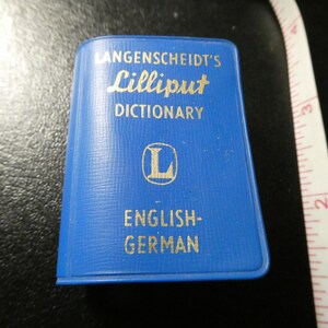 May include: A small, blue Langenscheidt's Lilliput Dictionary with gold lettering. The cover reads "ENGLISH-GERMAN". A white measuring tape with black markings is in the background.