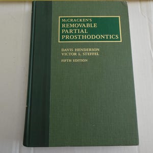 Puede incluir: Un libro de tapa dura verde titulado "McCracken's Removable Partial Prosthodontics". El título está en un recuadro rectangular dorado. Los nombres de los autores y el número de edición también son visibles.