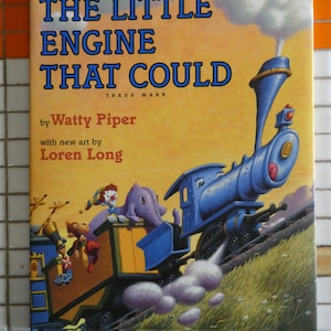 Könnte beinhalten: Ein Vintage-Kinderbuchcover mit einer blauen Lokomotive, die einen Wagen mit einem Clown, einer Giraffe und einem Elefanten zieht. Der Buchtitel lautet "The Little Engine That Could" von Watty Piper mit neuen Illustrationen von Loren Long.