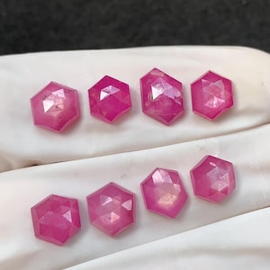 Pode incluir: Oito pedras preciosas rosas facetadas, em forma hexagonal. As pedras são de uma cor magenta vibrante e parecem ser cortadas com múltiplas facetas, refletindo a luz. As pedras preciosas são exibidas em uma superfície branca.