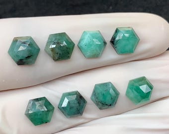 May include: Eight faceted emerald gemstones in a hexagonal shape. The gemstones vary in shades of green, with some showing darker inclusions. The gems are displayed on a white surface, suggesting they are ready for jewelry making.