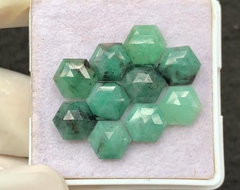 May include: A collection of faceted emerald gemstones in a hexagonal shape, arranged in a honeycomb pattern. The gems vary in shades of green, from deep forest to lighter hues, and are displayed in a white tray.
