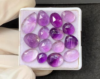 May include: A selection of faceted amethyst gemstones in various shapes and sizes, presented in a white tray. The gemstones range in colour from light lavender to deep purple, with visible internal patterns and cuts.