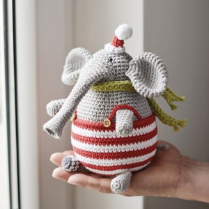 May include: A grey crocheted elephant wearing a red and white striped outfit, a green scarf, and a red Santa hat.