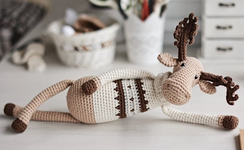 May include: A crocheted moose toy with brown antlers, a brown and white striped sweater, and brown hooves. The moose is lying on its side with its head turned to the right.