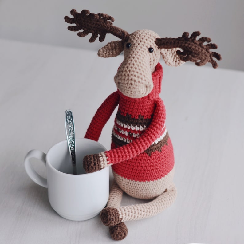 May include: A crocheted moose wearing a red sweater with white and brown stripes. The moose is sitting next to a white mug and a silver spoon.