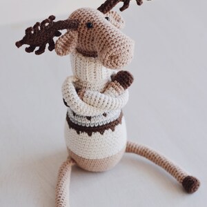 May include: A crocheted moose toy wearing a white sweater with brown and gray stripes. The moose has brown antlers and is sitting on a white surface.
