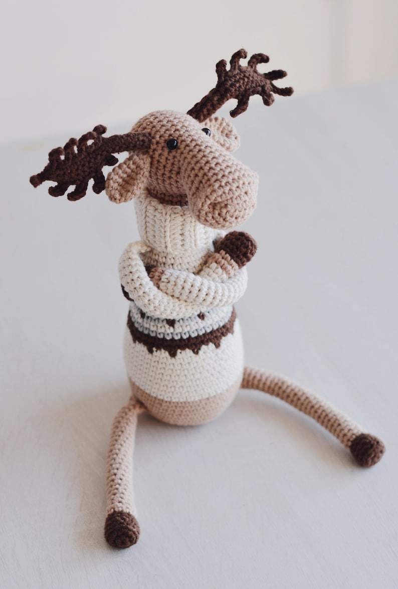 May include: A crocheted moose toy wearing a white sweater with brown and gray stripes. The moose has brown antlers and is sitting on a white surface.
