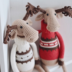 May include: Two crocheted moose toys wearing sweaters. One moose is wearing a white sweater with brown stripes and the other is wearing a red sweater with brown and white stripes.