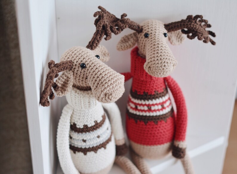 May include: Two crocheted moose toys wearing sweaters. One moose is wearing a white sweater with brown stripes and the other is wearing a red sweater with brown and white stripes.