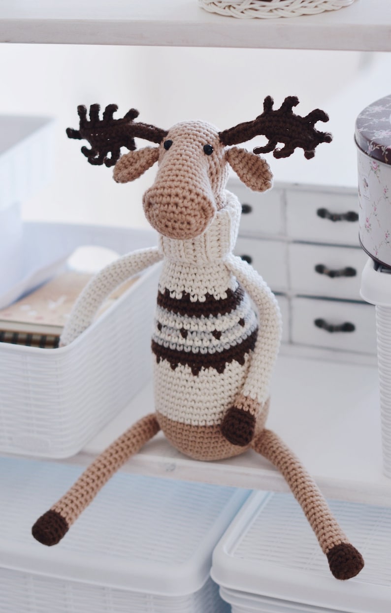 May include: A crocheted moose toy with brown antlers, a white sweater with brown and gray stripes, and a brown tail. The moose is sitting on a white shelf.