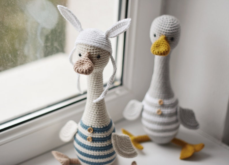 Peut inclure: Deux jouets en crochet en forme d'oie, l'un portant un chapeau de lapin blanc avec de longues oreilles et une chemise ray&eacute;e bleu et blanche, l'autre avec une chemise ray&eacute;e gris et blanche.
