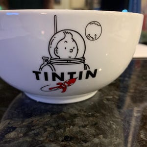 Może przedstawiać: Biała ceramiczna miska z czarno-czerwoną grafiką Tintina w skafandrze kosmicznym, rakietą i księżycem. Słowo "TINTIN" jest wydrukowane na czarno pod ilustracją. Miska ma zaokrąglony kształt.