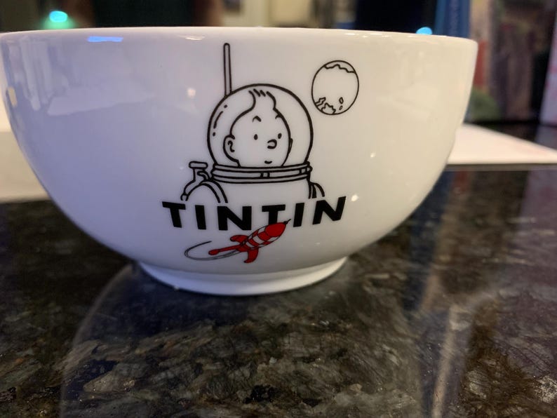 Może przedstawiać: Biała ceramiczna miska z czarno-czerwoną grafiką Tintina w skafandrze kosmicznym, rakietą i księżycem. Słowo "TINTIN" jest wydrukowane na czarno pod ilustracją. Miska ma zaokrąglony kształt.