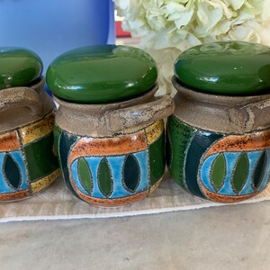 May include: Three ceramic jars with green lids and decorative patterns in blue, green, yellow, and orange. Each jar has a handle and a wooden spoon. The jars are on a white surface.