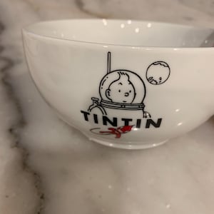 Może przedstawiać: Biała ceramiczna miska z czarno-białą ilustracją Tintina w skafandrze kosmicznym i napisem "TINTIN" poniżej. Miska ma zaokrąglony kształt i gładkie, błyszczące wykończenie. Widoczny jest mały czerwony akcent.