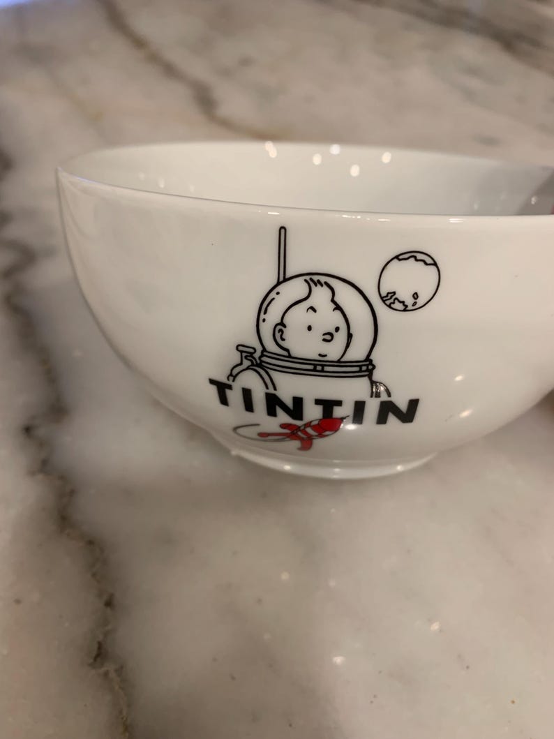 Może przedstawiać: Biała ceramiczna miska z czarno-białą ilustracją Tintina w skafandrze kosmicznym i napisem "TINTIN" poniżej. Miska ma zaokrąglony kształt i gładkie, błyszczące wykończenie. Widoczny jest mały czerwony akcent.