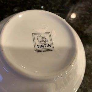 Może przedstawiać: Biała ceramiczna miska z logo Tintina na spodzie. Logo przedstawia Tintina i Milusia w kwadratowej ramce. Miska ma lekko zakrzywiony kształt i błyszczące wykończenie. Prawdopodobnie przedmiot kolekcjonerski.